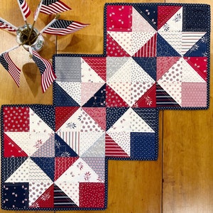 May include: Three patriotic quilted placemats in red, white, and blue, arranged on a wooden surface. Each square placemat features a patchwork design with various patterns and a dark blue border. Small American flags are in a vase.