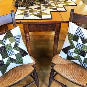 May include: Two wooden chairs with woven seats and green and white patchwork pillows. The chairs are set at a wooden table with a green and white patchwork table runner.