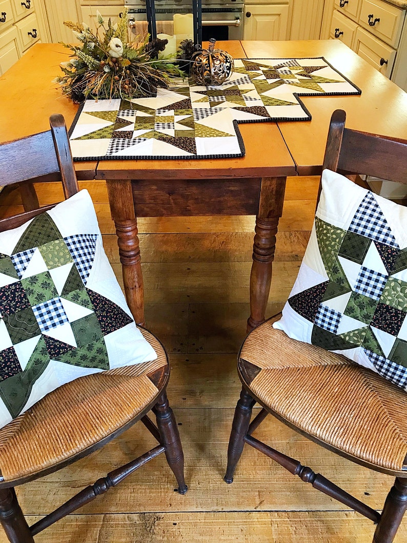 May include: Two wooden chairs with woven seats and green and white patchwork pillows. The chairs are set at a wooden table with a green and white patchwork table runner.
