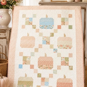 May include: A rectangular quilt featuring a white background with a repeating pattern of five pieced fabric pumpkins in shades of blue, peach, and green. The quilt is framed by a peach-coloured floral border and displays intricate stitching.