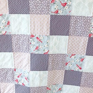 May include: A patchwork quilt with a variety of patterns, including floral, geometric, and polka dot designs. The quilt is made in shades of gray, beige, and light blue, with red birds featured on some of the squares.