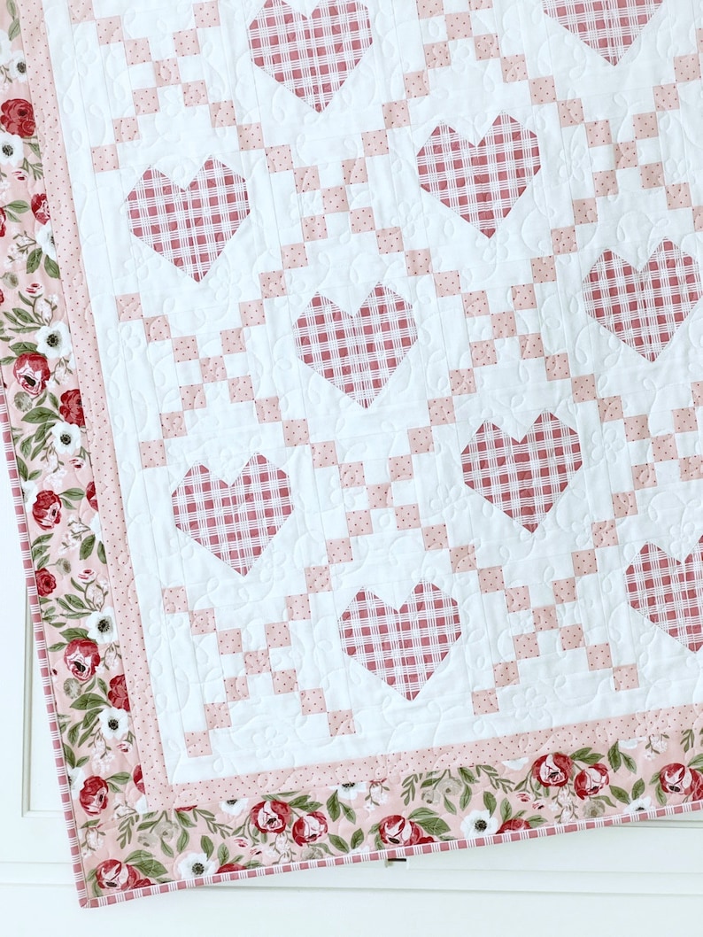 May include: A white quilt with a pink and white gingham pattern in the shape of hearts. The quilt has a floral border with pink, white, and green flowers.