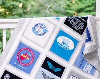 May include: A quilt made from t-shirts with various designs and text. The quilt is made up of squares with a white background and a gray border. The designs include a horse, a state logo, a bird, and a graduation design. The text includes "COLTS", "Virginia Governer's Honers Academy", "SOLS", "GHLA", "Fairmont State University", "The Sustainable Life", "Caring-oms", "how", "enristians", "f the salt", "re the salt of the earth", "salt loses it's saltiness", "made salty again?", "It is no longer goo", "except to be thrown", "trampled underfoot."