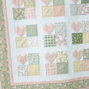 K&ouml;nnte beinhalten: Eine wei&szlig;e Steppdecke mit einem rosafarbenen und gr&uuml;nen Patchwork-Muster. Die Steppdecke besteht aus Quadraten mit Herzen in der Mitte. Die Steppdecke hat einen rosafarbenen und gr&uuml;nen Rand.