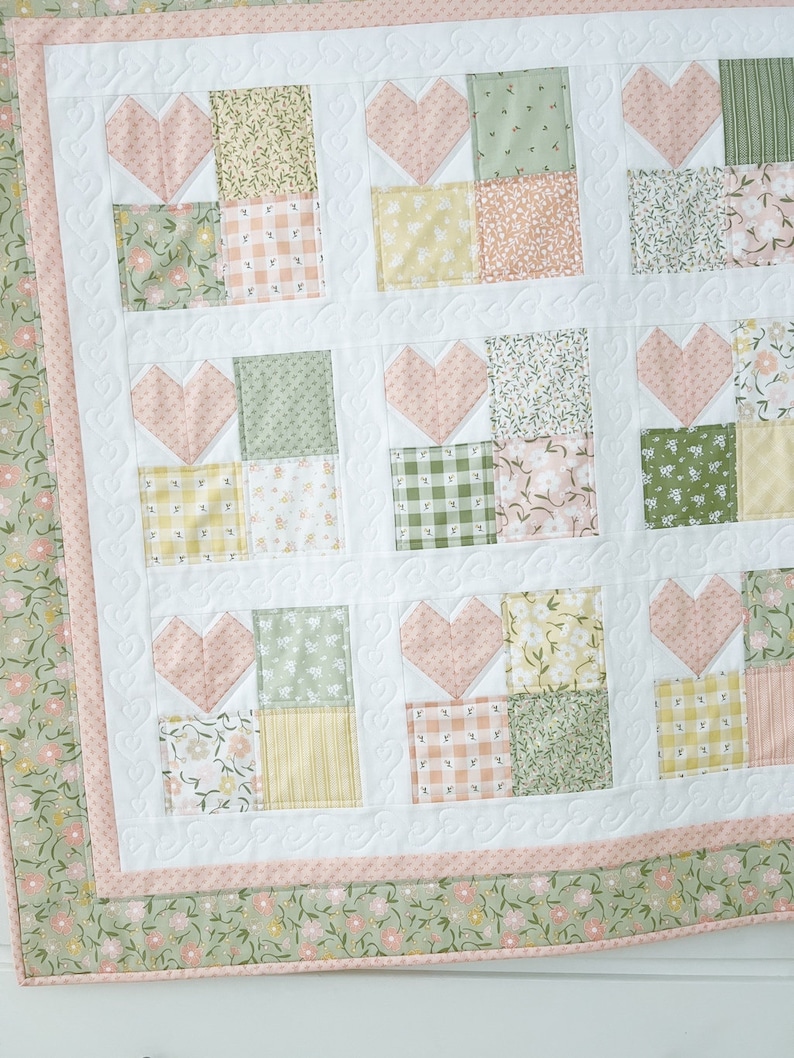 K&ouml;nnte beinhalten: Eine wei&szlig;e Steppdecke mit einem rosafarbenen und gr&uuml;nen Patchwork-Muster. Die Steppdecke besteht aus Quadraten mit Herzen in der Mitte. Die Steppdecke hat einen rosafarbenen und gr&uuml;nen Rand.