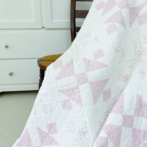 Peut inclure: Une courtepointe blanche et rose avec un motif floral est drap&eacute; sur une chaise en bois. La courtepointe est faite avec un design de patchwork de carr&eacute;s et de triangles.