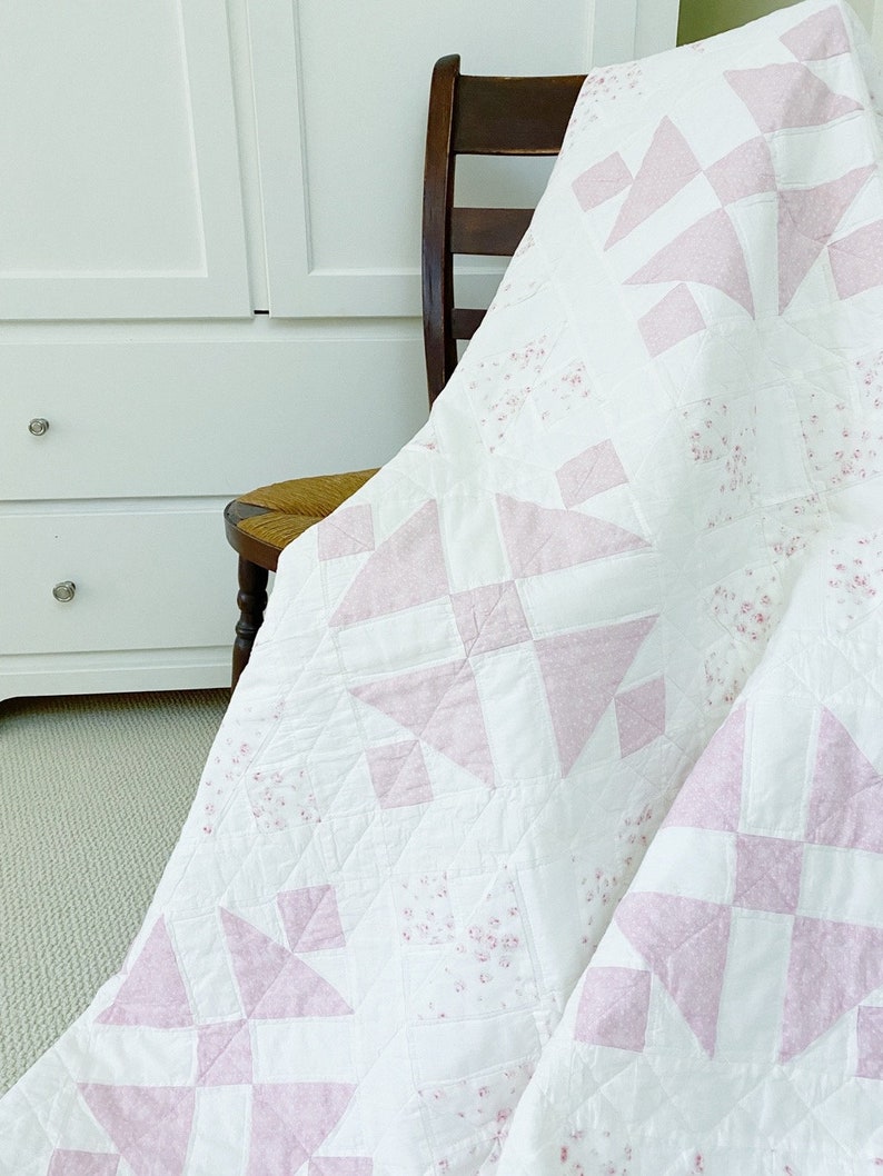 Peut inclure: Une courtepointe blanche et rose avec un motif floral est drap&eacute; sur une chaise en bois. La courtepointe est faite avec un design de patchwork de carr&eacute;s et de triangles.