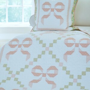 May include: A white quilt and pillow set with a repeating bow pattern in peach and white polka dots. The quilt features a green floral border and a matching pillow with a peach bow design. The set is on a bed near a window.
