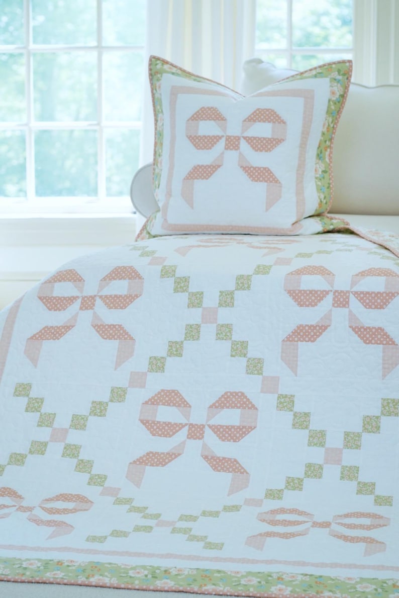 May include: A white quilt and pillow set with a repeating bow pattern in peach and white polka dots. The quilt features a green floral border and a matching pillow with a peach bow design. The set is on a bed near a window.