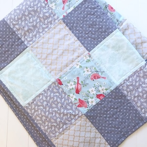 May include: A patchwork quilt with a gray, white, and blue color scheme. The quilt is made of different fabrics with floral and geometric patterns. The quilt is quilted with a decorative stitch.