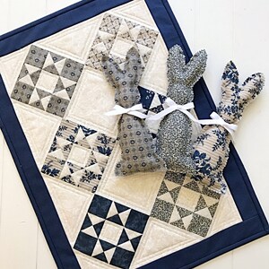May include: A blue and white patchwork quilt with a navy blue border. Two fabric bunny toys are placed on top of the quilt.