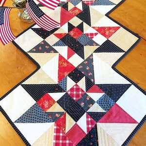 Puede incluir: Un camino de mesa acolchado rojo, blanco y azul con un patrón de estrellas. El camino de mesa está hecho de varias telas con diferentes patrones, incluyendo cuadros, flores y lunares. El camino de mesa está bordeado con un ribete negro.