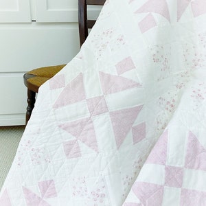 Peut inclure: Une courtepointe blanche avec un motif floral rose et blanc. La courtepointe est pli&eacute;e et drap&eacute;e sur une chaise en bois.