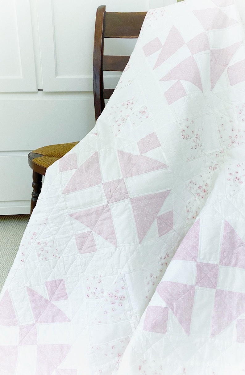Peut inclure: Une courtepointe blanche avec un motif floral rose et blanc. La courtepointe est pli&eacute;e et drap&eacute;e sur une chaise en bois.