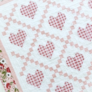 May include: A white quilt with a pink and white gingham pattern. The quilt is decorated with red hearts with a gingham pattern. The quilt has a pink border with a floral pattern.