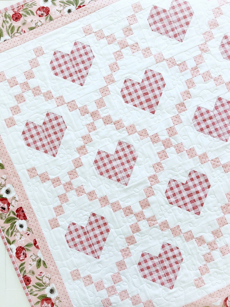 May include: A white quilt with a pink and white gingham pattern. The quilt is decorated with red hearts with a gingham pattern. The quilt has a pink border with a floral pattern.