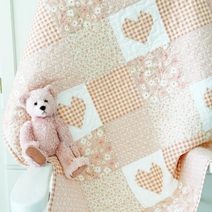 May include: A pink and white patchwork quilt with a gingham pattern and floral print. The quilt is folded and draped over a white chair with a pink teddy bear sitting on it.