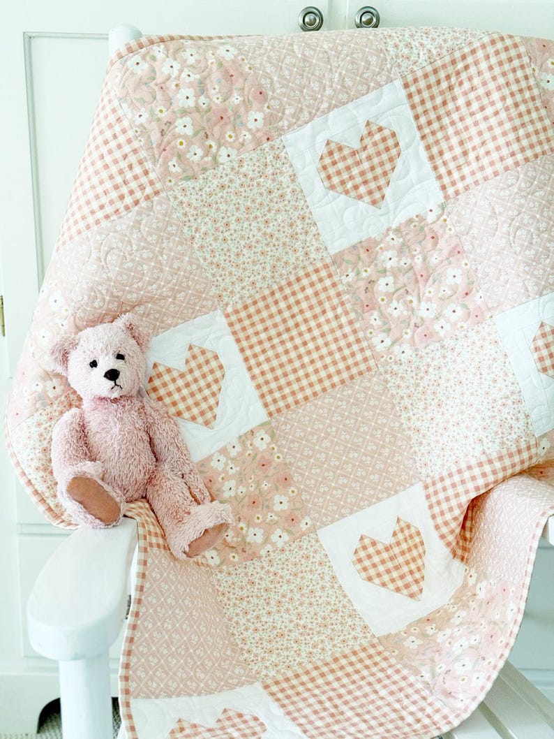 May include: A pink and white patchwork quilt with a gingham pattern and floral print. The quilt is folded and draped over a white chair with a pink teddy bear sitting on it.