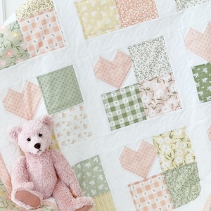 May include: A pink and green patchwork quilt with a heart pattern. A pink teddy bear is sitting on the quilt.