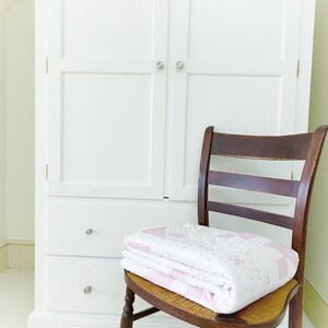 Peut inclure: Une armoire en bois blanc avec deux portes et deux tiroirs. Une chaise en bois avec un si&egrave;ge en osier est devant l'armoire. Une courtepointe rose et blanche est pli&eacute;e sur le si&egrave;ge de la chaise.