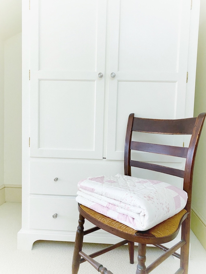 Peut inclure: Une armoire en bois blanc avec deux portes et deux tiroirs. Une chaise en bois avec un si&egrave;ge en osier est devant l'armoire. Une courtepointe rose et blanche est pli&eacute;e sur le si&egrave;ge de la chaise.