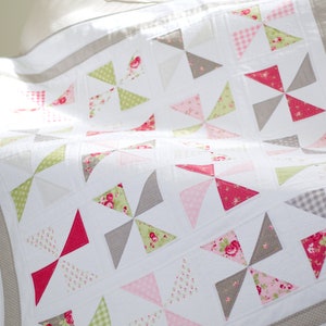 Peut inclure: Une courtepointe blanche avec un motif de moulinet. Les moulinets sont faits de tissus à motifs floraux roses, verts et gris. La courtepointe a une bordure blanche et une bordure marron clair.