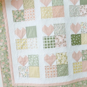 K&ouml;nnte beinhalten: Eine rosafarbene und gr&uuml;ne Patchwork-Decke mit einem Herz-Design. Die Decke besteht aus verschiedenen floralen und karierten Stoffen.