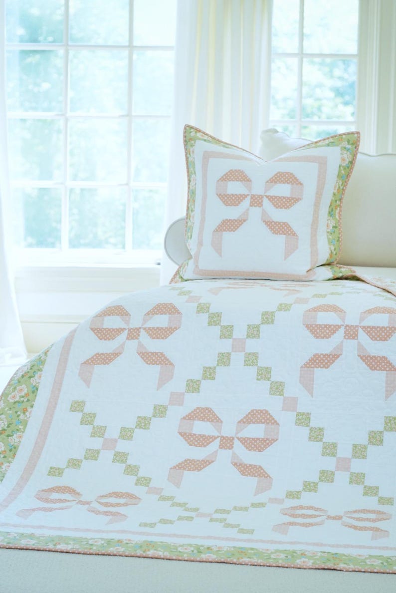 May include: A white quilt and pillow set featuring a repeating bow pattern in peach and white polka dots. The quilt has a floral border in shades of green and peach. The pillow is on a bed in front of a window.