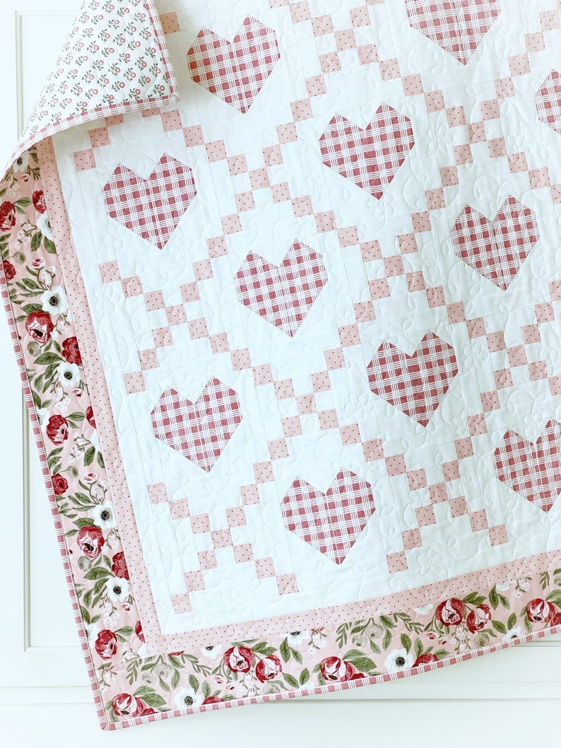 May include: A white quilt with a pink and white gingham pattern. The quilt is decorated with pink hearts and has a floral border.