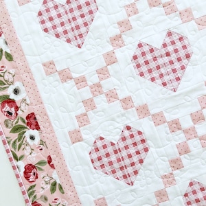 May include: A white quilt with a pink and white gingham pattern. The quilt has three pink hearts with a gingham pattern in the center of each heart. The quilt has a pink border with a floral pattern.