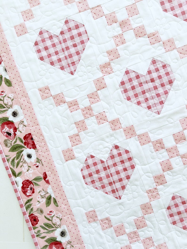 May include: A white quilt with a pink and white gingham pattern. The quilt has three pink hearts with a gingham pattern in the center of each heart. The quilt has a pink border with a floral pattern.