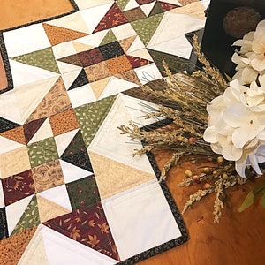 May include: A quilted table runner with a patchwork design featuring a variety of fall colors, including brown, green, cream, and red. The runner has a black border and is displayed on a wooden table.