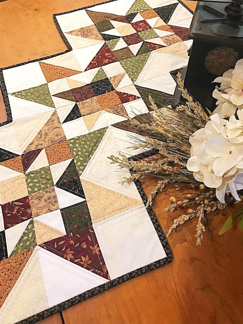 May include: A quilted table runner with a patchwork design featuring a variety of fall colors, including brown, green, cream, and red. The runner has a black border and is displayed on a wooden table.