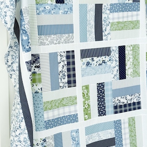 May include: A patchwork quilt in shades of blue, green, and white. The quilt's design is a grid of rectangular blocks, each with a different fabric pattern, including floral, striped, and dotted designs. The border is white with a blue floral pattern.