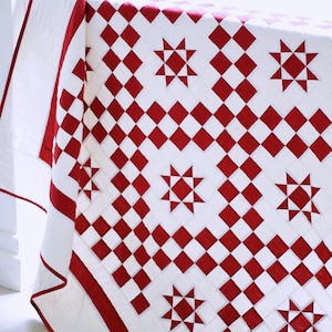 May include: A white and red quilt with a starlit path design. The quilt is folded and draped over a white surface. The text "Starlit Path by Maple Cottage Designs" is visible at the top of the image.