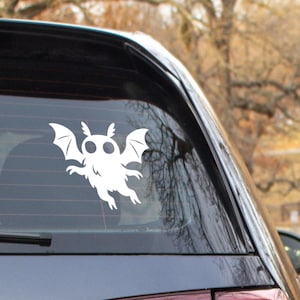 May include: A white vinyl decal of a cartoonish bat-like creature with large eyes and wings, affixed to the rear window of a vehicle. The design is simple and graphic, suitable for Halloween or fantasy themes.