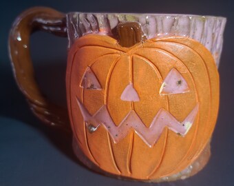 May include: A ceramic mug shaped like a jack-o'-lantern with a pink and brown glaze. The mug has a handle and a wide opening.
