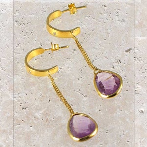 May include: A pair of gold hoop earrings with a dangling amethyst gemstone. The hoops are accented with small clear stones.