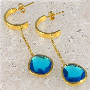 Puede incluir: Aretes de aro de oro con una piedra preciosa azul colgante. Los aretes tienen una cadena delicada que conecta el aro a la piedra preciosa.