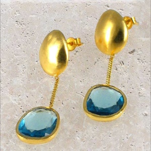 Può includere: Un paio di orecchini d'oro con gocce di pietre preziose blu. Gli orecchini presentano una parte superiore liscia di forma ovale e una pietra preziosa blu pendente.