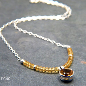 May include: A silver chain necklace with a citrine gemstone pendant. The necklace is made with small, round citrine beads and a larger, round citrine gemstone in the centre. The necklace is on a grey background.