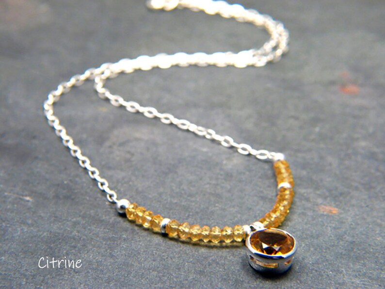 May include: A silver chain necklace with a citrine gemstone pendant. The necklace is made with small, round citrine beads and a larger, round citrine gemstone in the centre. The necklace is on a grey background.