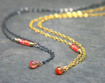 May include: Two delicate necklaces with gemstone pendants. One is gold-toned, the other dark grey. Both feature small red gemstone beads and teardrop pendants. The chains are fine and elegant.