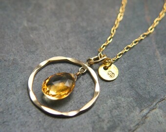 May include: A gold necklace featuring a hammered circle pendant with a teardrop-shaped citrine gemstone. The necklace has a small gold tag with the letter "B" on it.