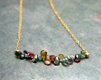 May include: A gold chain necklace featuring a cluster of colourful gemstones in a rainbow pattern. The gemstones are teardrop shaped and include green, blue, pink, and brown.