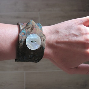 Può includere: Un braccialetto in tessuto con motivo marrone e blu con un bottone bianco. Il bottone ha due fori.