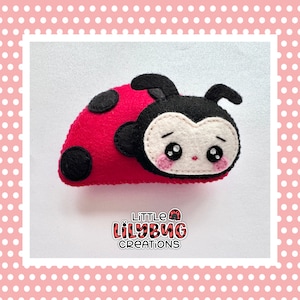 Puede incluir: Una mariquita de fieltro roja y negra con cara blanca y mejillas rosadas. La mariquita está tumbada de espaldas con las patas en el aire. El texto "LITTLE LILYBUG CREATIONS" está impreso debajo de la mariquita.