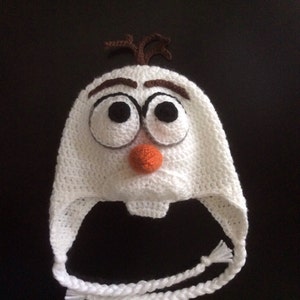 May include: A white crocheted hat in the style of Olaf from Frozen. It has large black eyes, brown eyebrows, an orange nose, and brown hair. White braided ties are attached to the ear flaps. A fun, handmade winter accessory.