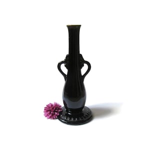 May include: A black vase with a round base and two handles. The vase has a tall, slender neck and a decorative rim. A purple flower rests near the base of the vase. The vase is made of a glossy material.