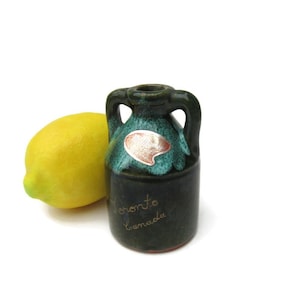 May include: A small, dark green ceramic jug with two handles and a turquoise top, next to a yellow lemon. The jug has a label and the words "Toronto Canada" written on it.